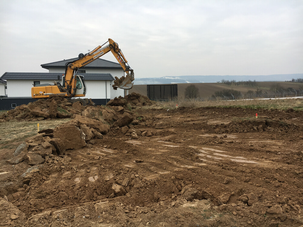 Bagger gräbt Baugrube auf freiem Grundstück; Erdehaufen und Wohnhaus im Hintergrund