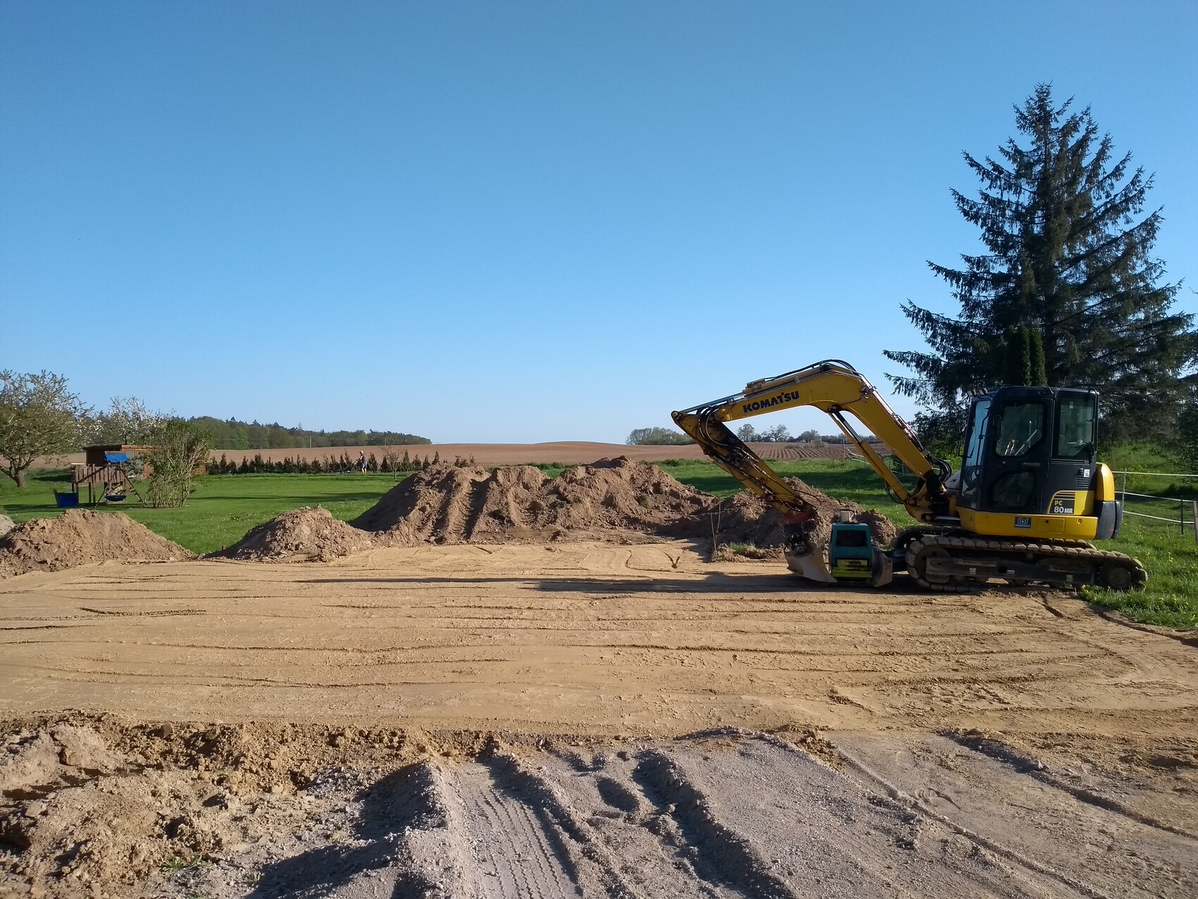 Gelber Komatsu-Bagger gräbt Erde auf Baufläche; Sandhügel, grüne Wiesen und Bäume im Hintergrund.