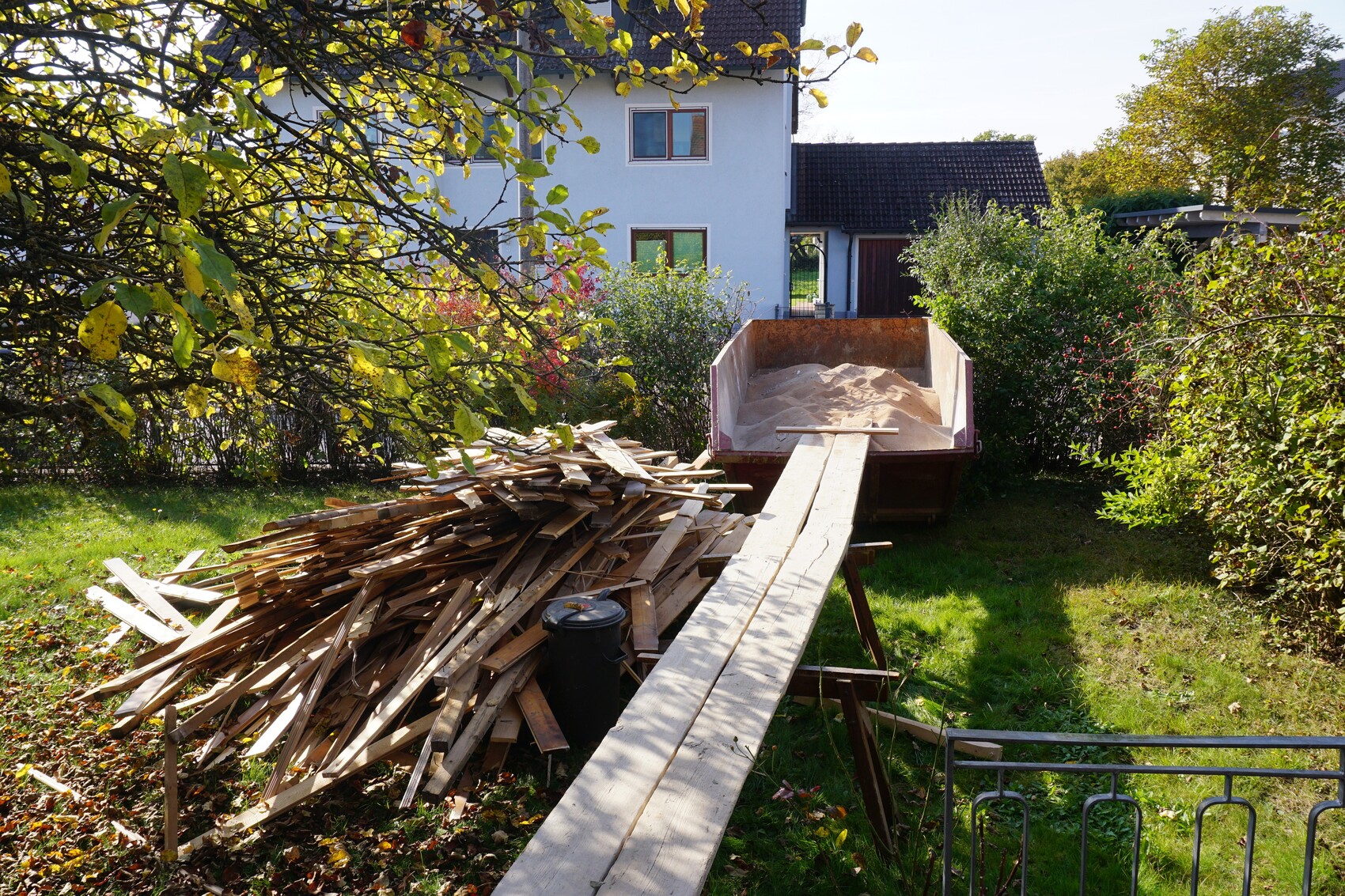 Gartenbaustelle: Holzlattenstapel, Rampe führt zu Sand im Anhänger, Haus im Hintergrund.