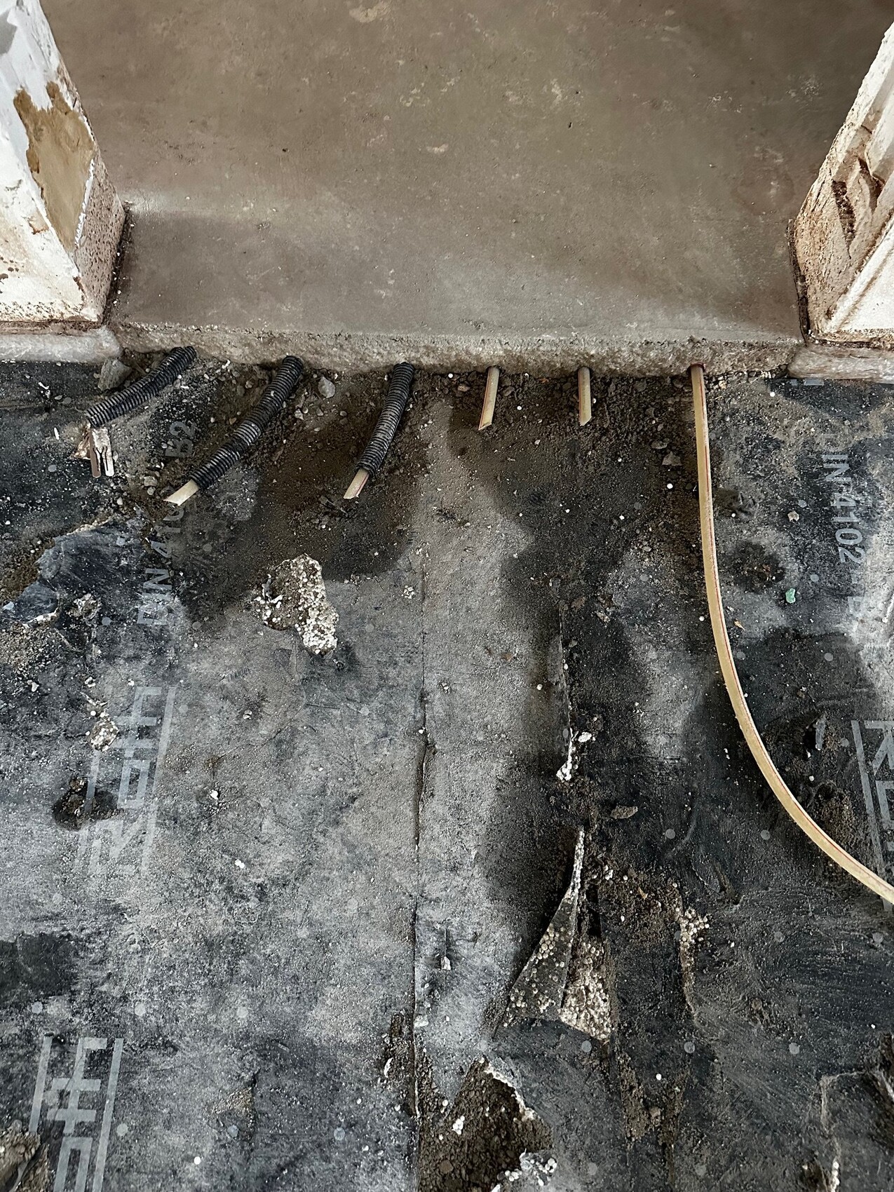 Baustellenboden mit schwarzen Kabelrohren, Holzstiften und losem Dämmmaterial nahe einer Türöffnung.