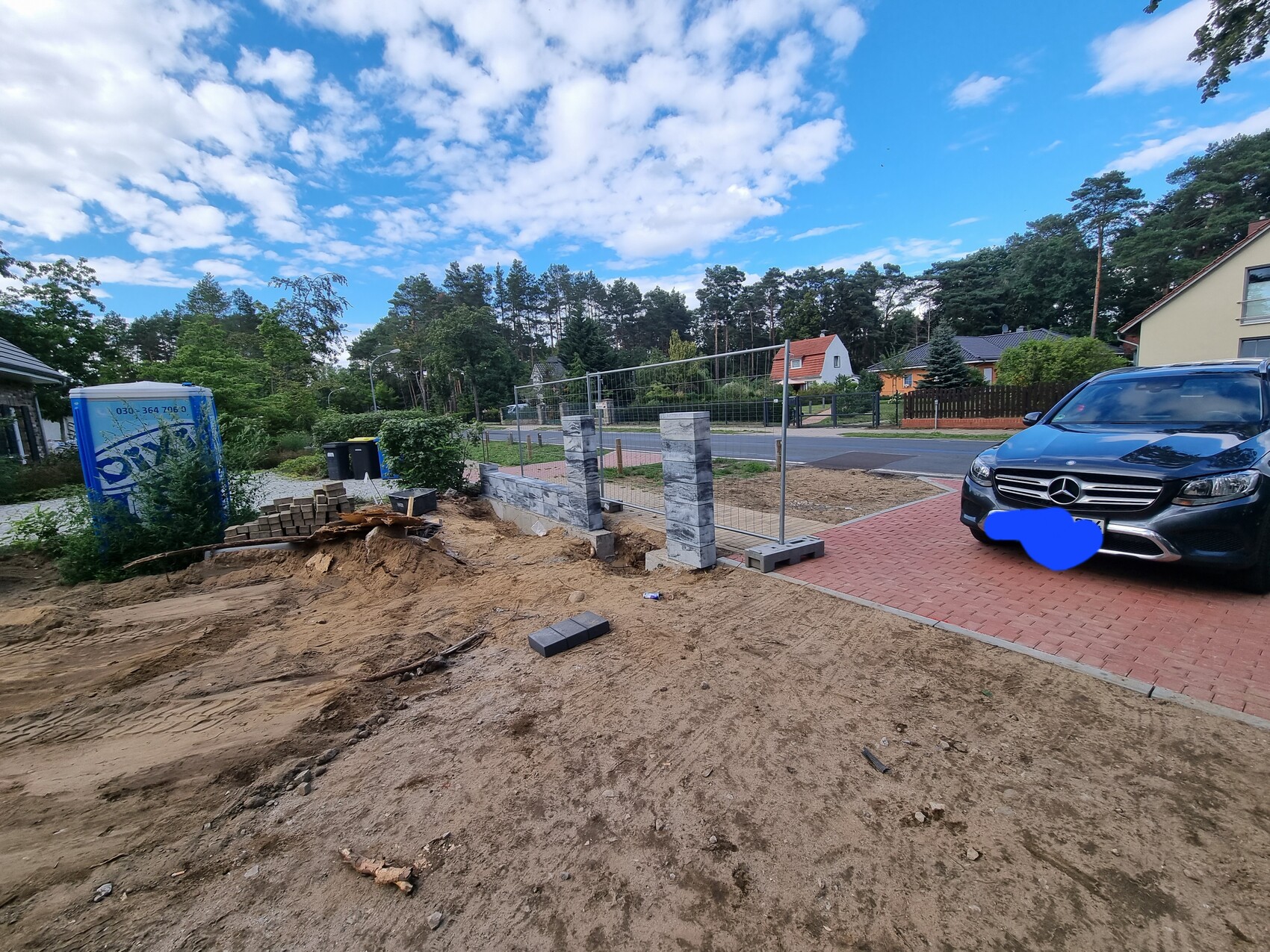 Baustelle im Vorgarten: Sand, Ziegel, Zaunpfosten; blaue Bau-Toilette, geparkter blauer Mercedes.