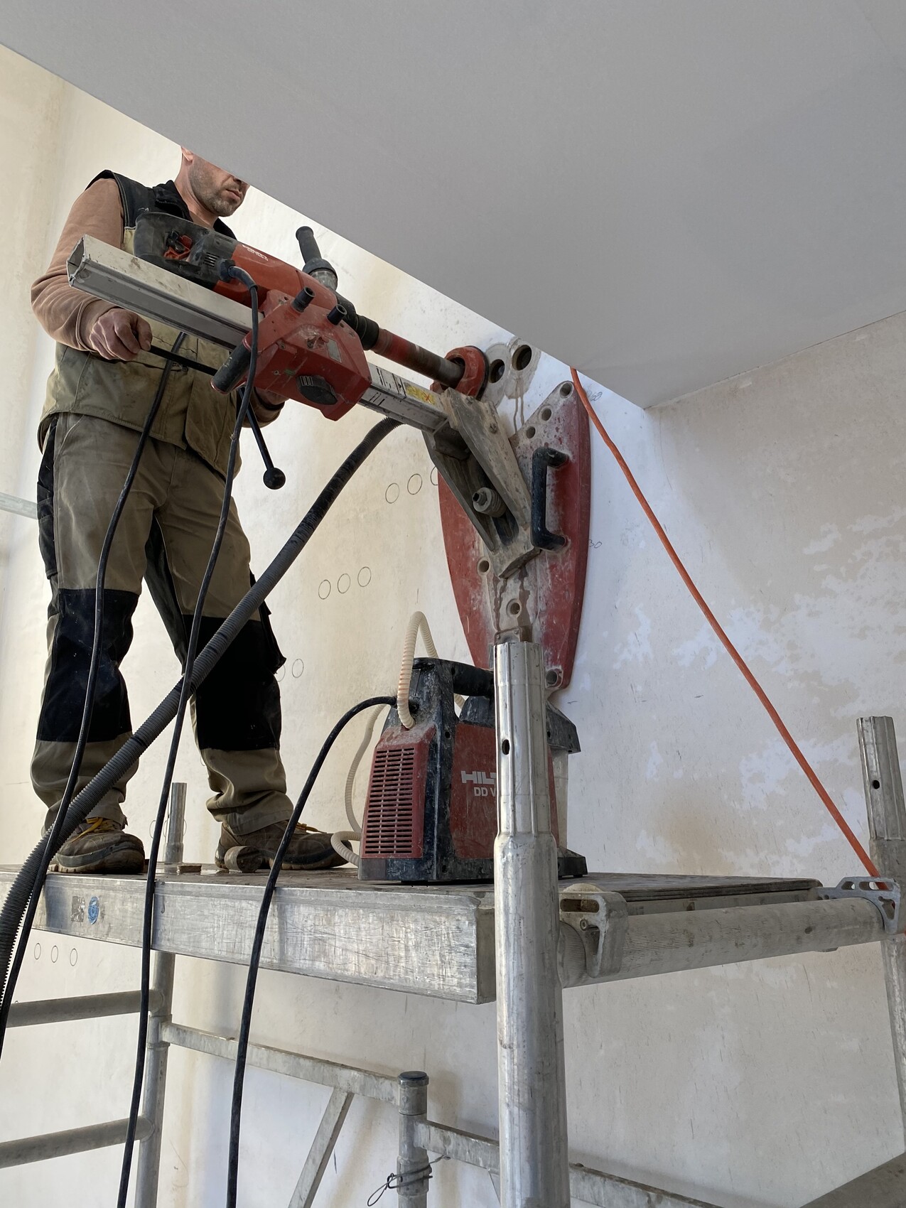 Arbeiter auf Gerüst bedient rote Wandbohrmaschine an Betonwand; Staubabsaugung sichtbar.