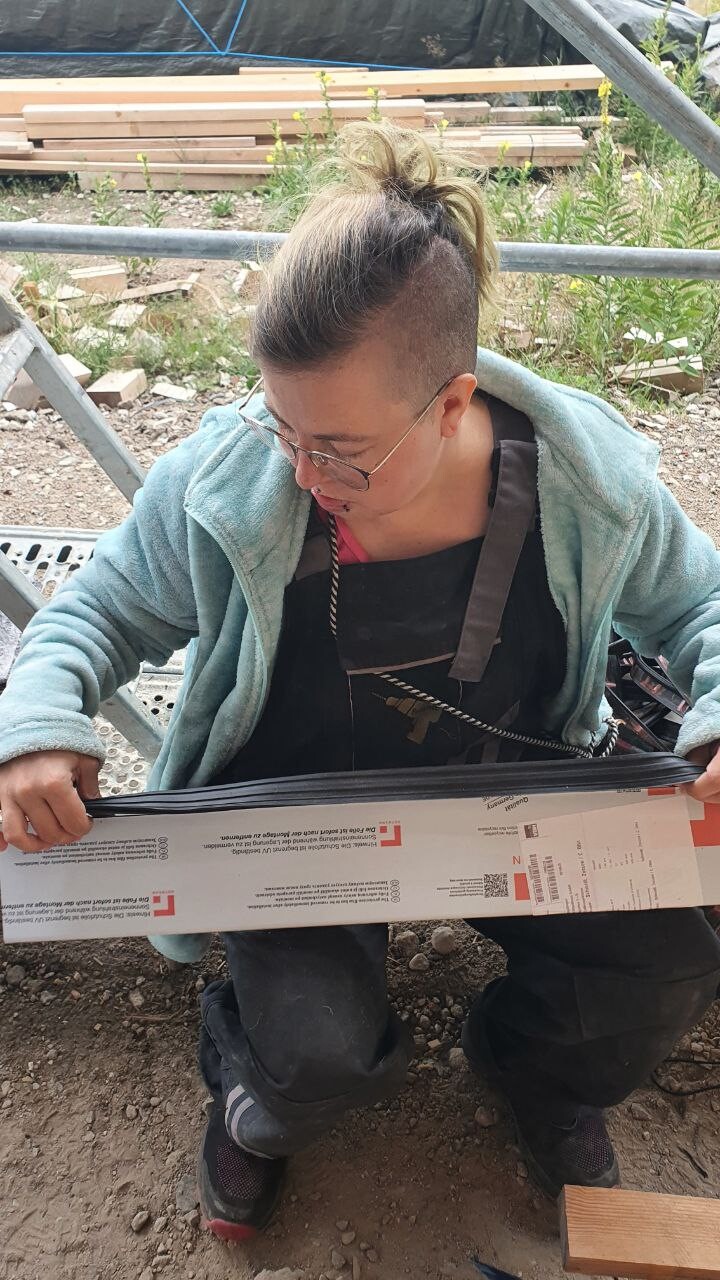 Person mit rasierten Seitenhaaren und Brille kniet auf Baustelle und hält einen Karton.