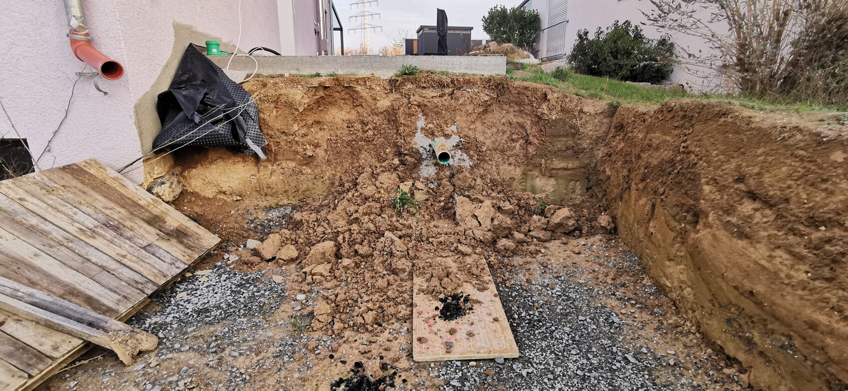 Große Baugrube im Garten neben dem Haus; lose Erde, Holzplatten und grüne Rohrleitung