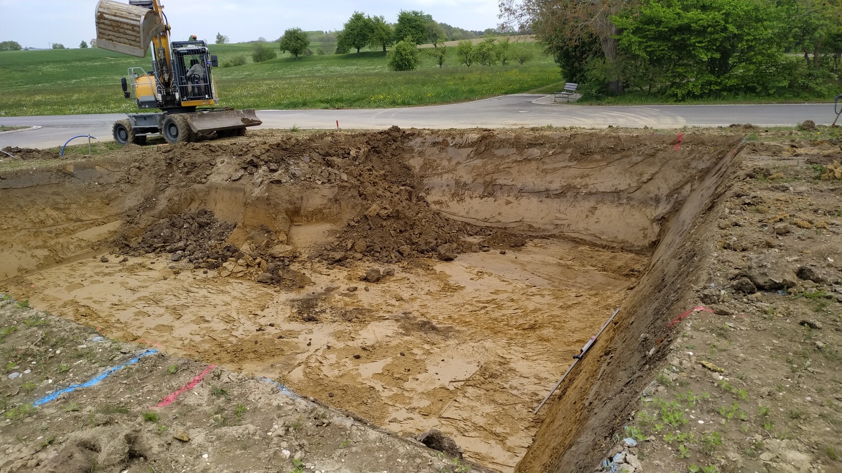 Großes Erdloch mit Schlamm, Bagger am Rand; Baugrube neben einer Straße und Grünland.
