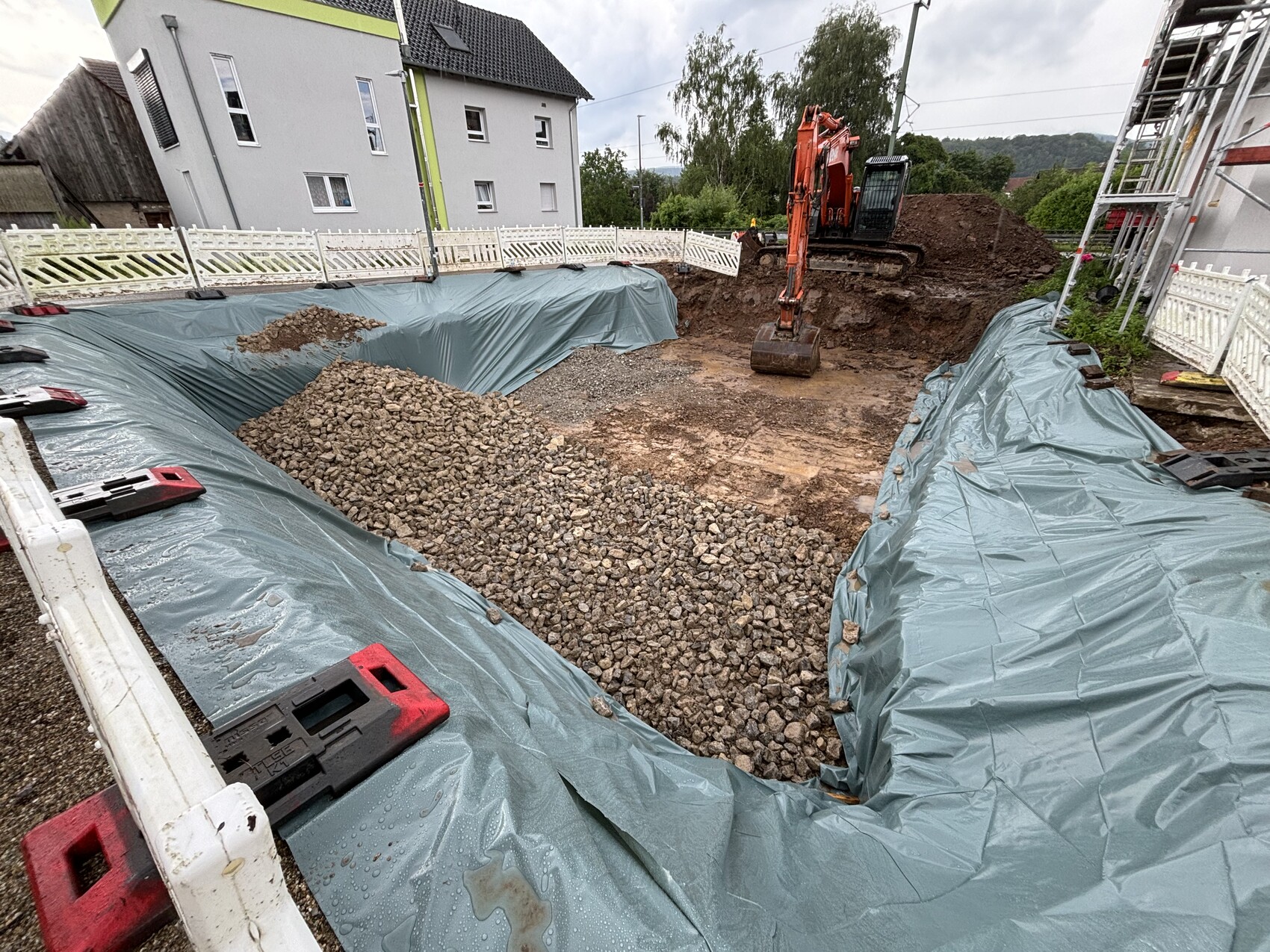 Baugrube mit blauer Plastikabdeckung, Kieshaufen und Bagger neben Häusern.
