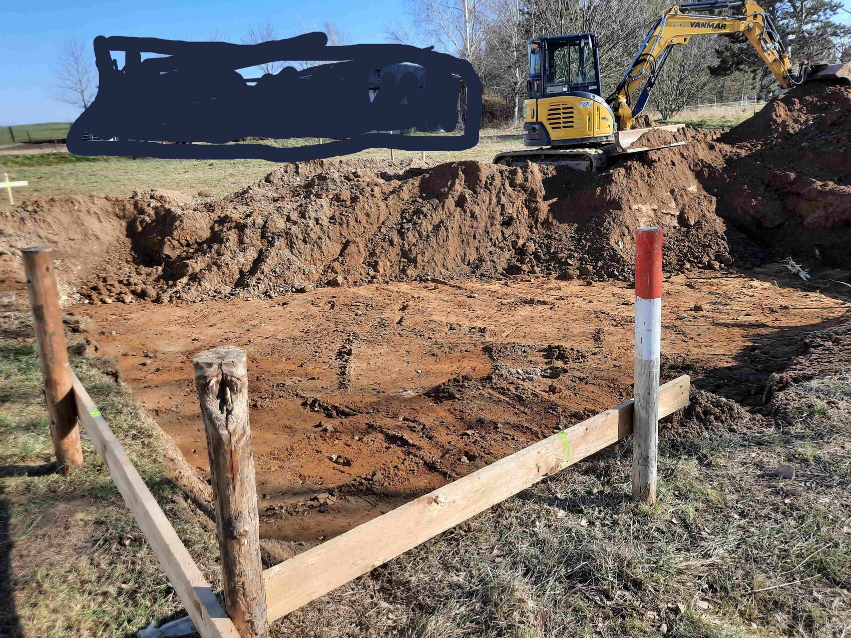 Baugrube auf Baustelle, Bagger im Hintergrund, Pfosten im Vordergrund.