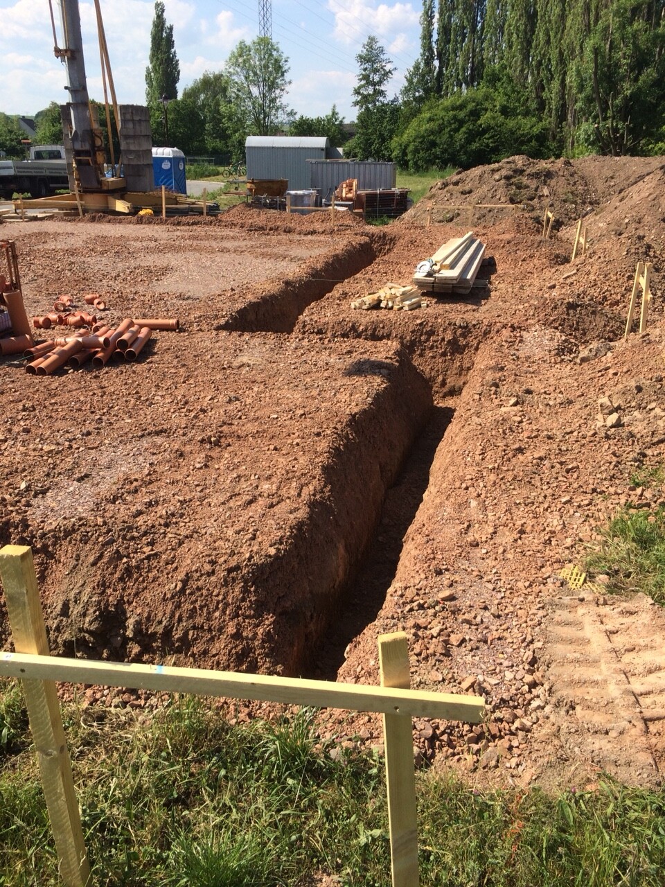 Baustelle mit tiefer Grube, links Rohre, rechts Holzstapel, Baugerät im Hintergrund