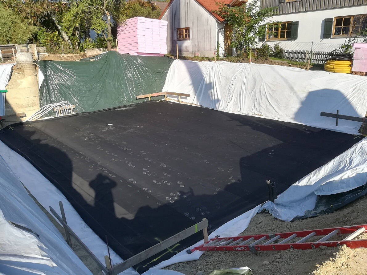 Fundament mit schwarzer Abdichtungsfolie, von Planen umgeben, auf Baustelle mit Materialien.