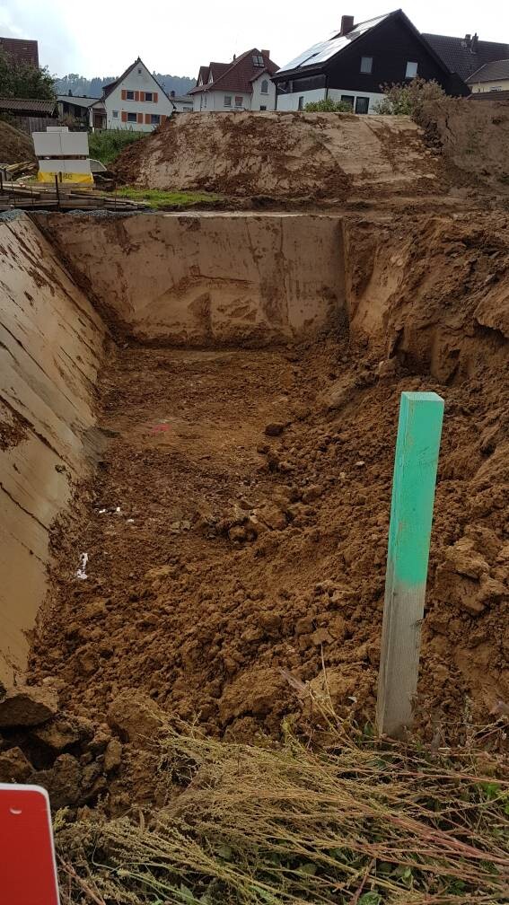 Ausgehobene Baugrube mit Erdreichwänden, grün markierter Pfosten, Häuser im Hintergrund.