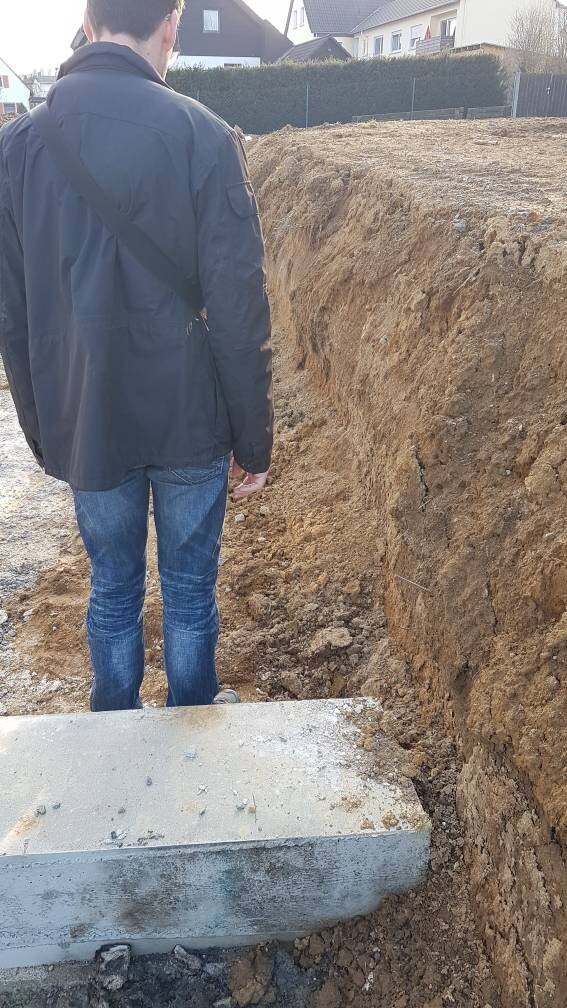 Person steht an einem tiefen Baugraben; am Rand liegt eine Betonplatte.