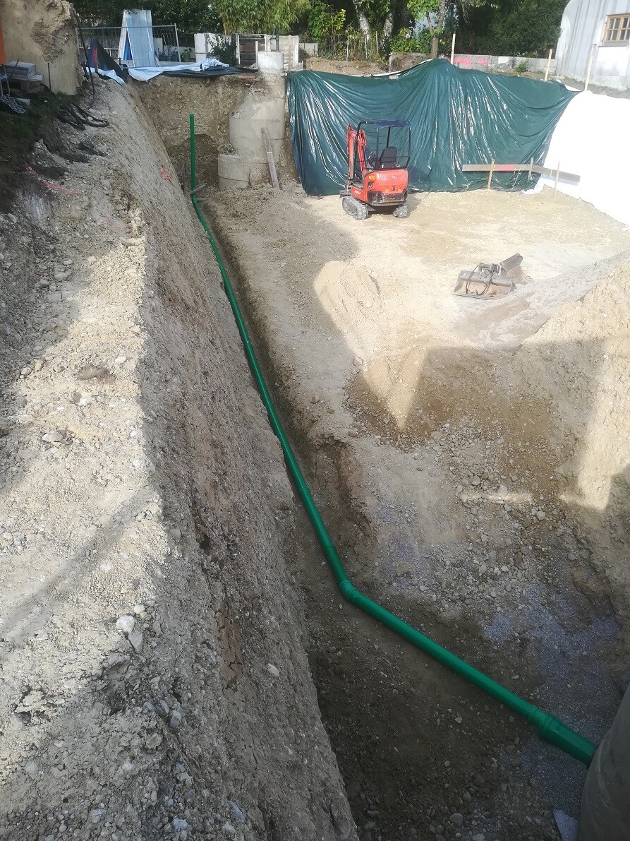 Tiefe Baugrube mit grünem Rohr entlang der Wand, Baugerät und Zelt im Hintergrund.