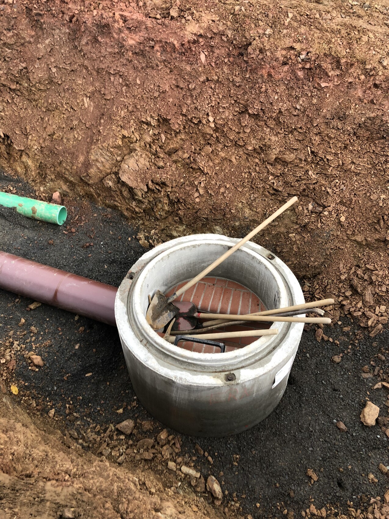 Runder Betonkanaldeckel im Graben mit Ziegelboden, Holzstöcken und Rohre daneben.