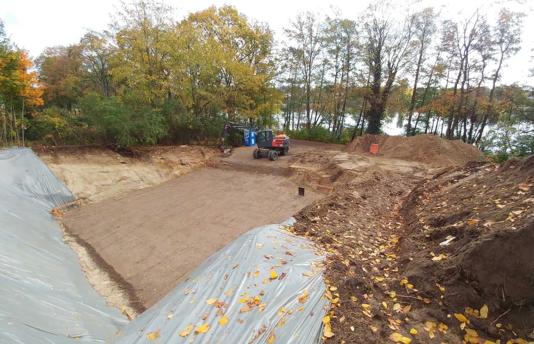 Baugrube im Wald: Bagger, Planierarbeiten, Erdaushub und Herbstlaub, Wald am Ufer im Hintergrund.
