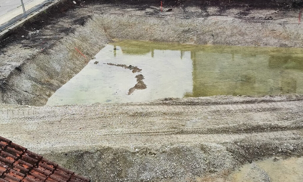 Ausgehobene Baugrube mit stehendem Wasserbecken, Kiesboden und Bauzaun.