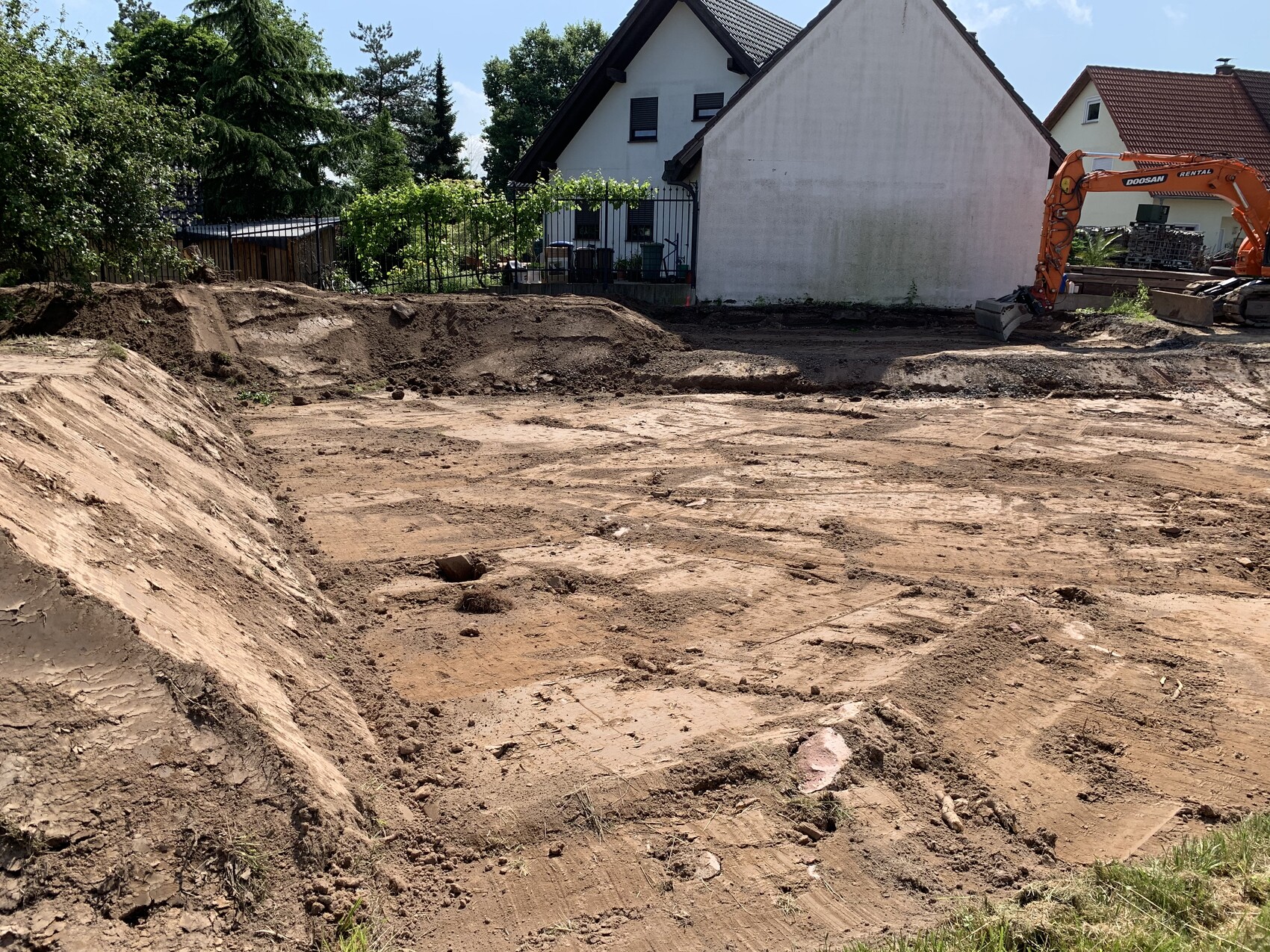 Baugrube mit ausgehobener Erde, orange Bagger rechts, Wohnhaus im Hintergrund