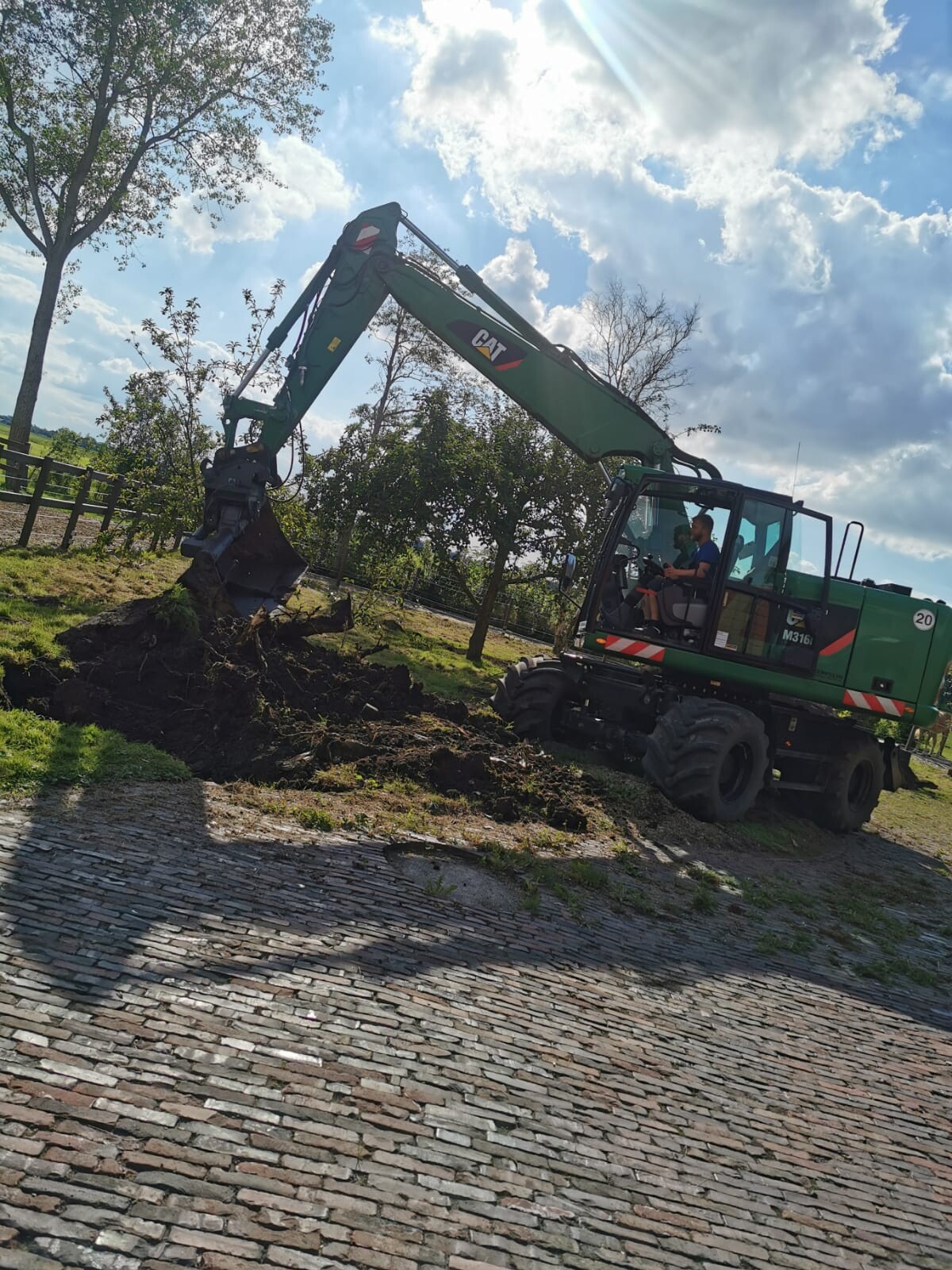 Grüner CAT-Bagger mit langem Arm gräbt Erde neben einem Kopfsteinpflasterweg; Fahrer im Fahrerhaus.