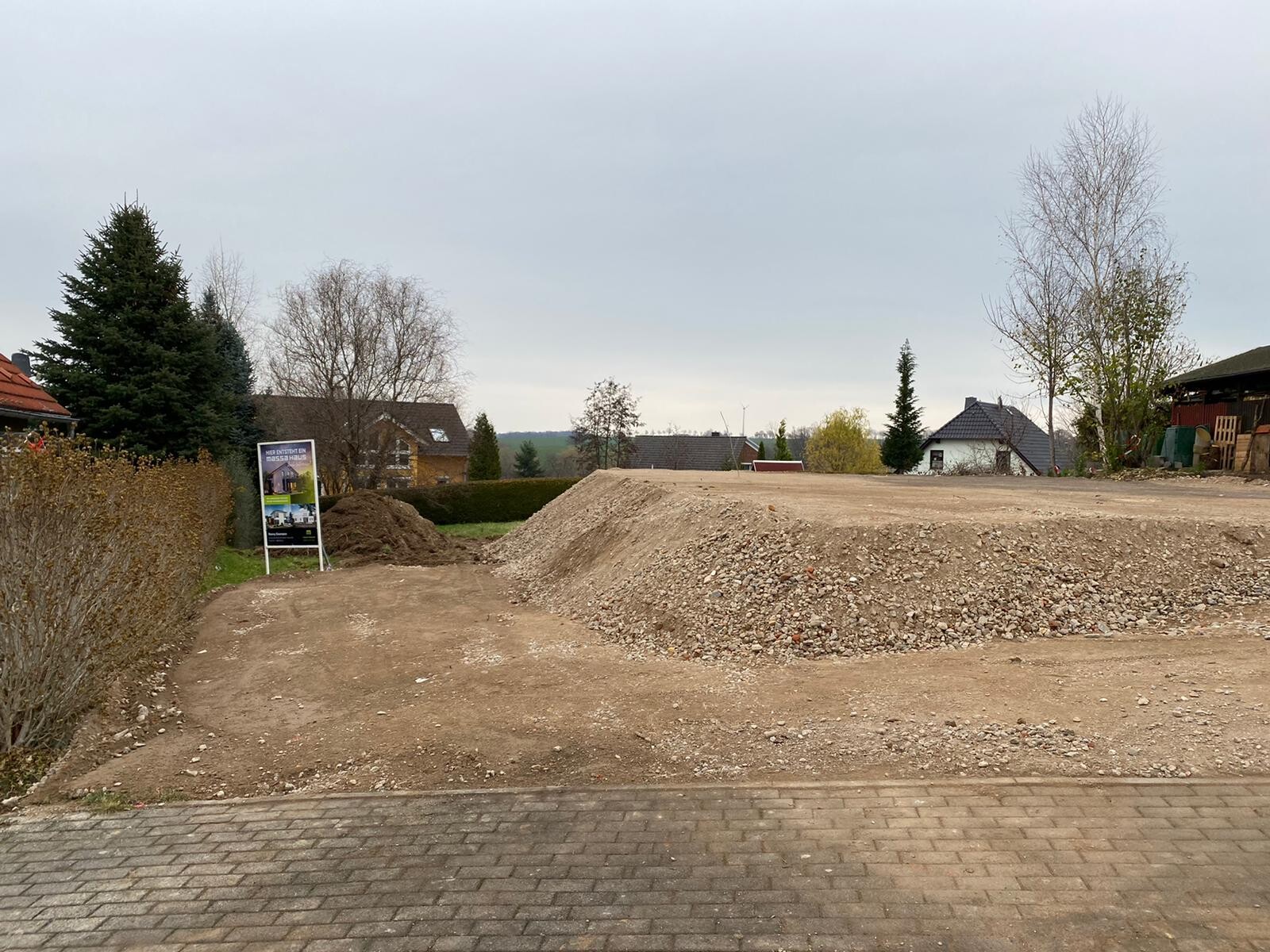 Baugrundstück mit Erdhügel, Kies, links Werbetafel, Vordergrund gepflastert, Häuser im Hintergrund