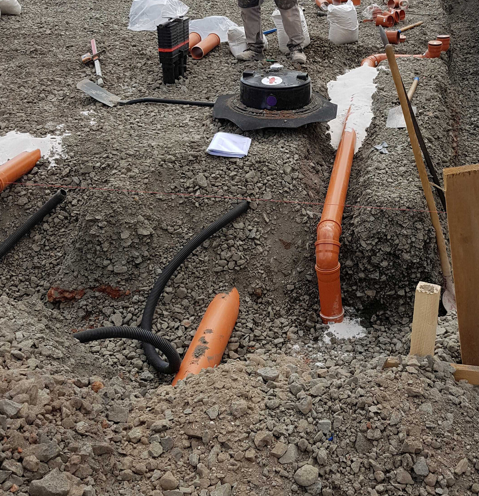 Baugrube mit orange PVC-Rohren, schwarzem Schlauch, Kiesuntergrund und Arbeitern im Hintergrund.