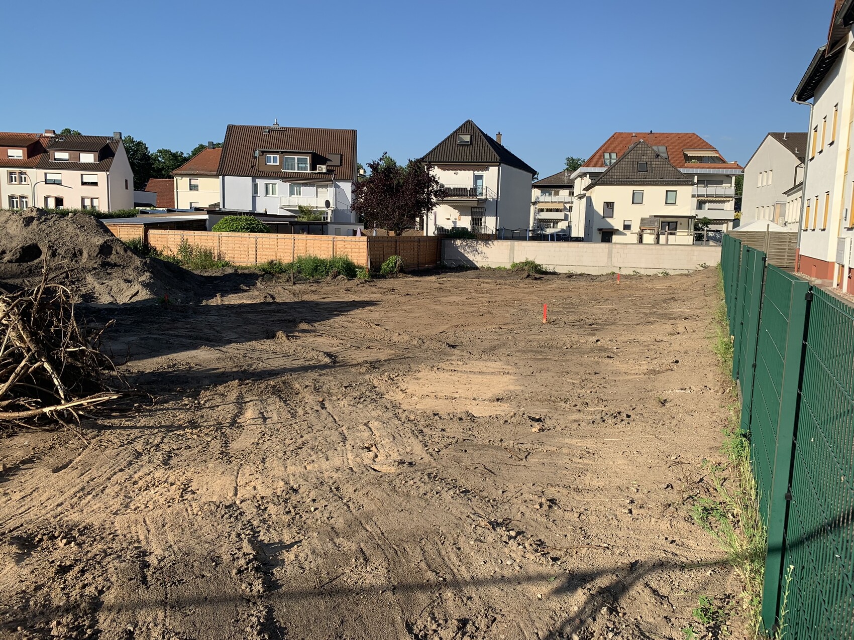 Leeres Baugrundstück mit Erdarbeiten, Zaun am Rand, Häuser im Hintergrund und blauem Himmel