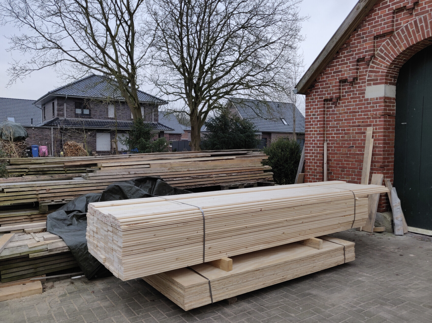 Gepackte Bauholzlattenstapel vor Backsteingebäude; Baum und weitere Holzbündel im Hintergrund.