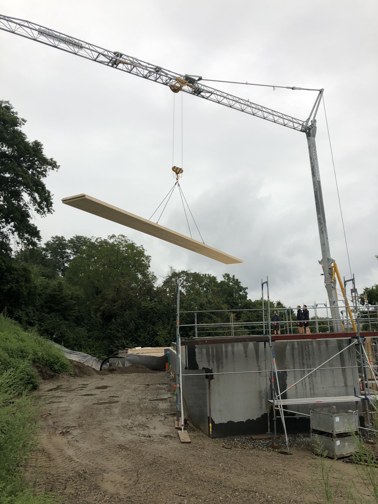 Baukran hebt einen langen Holzbalken über eine Baustelle mit Gerüst und Arbeitern
