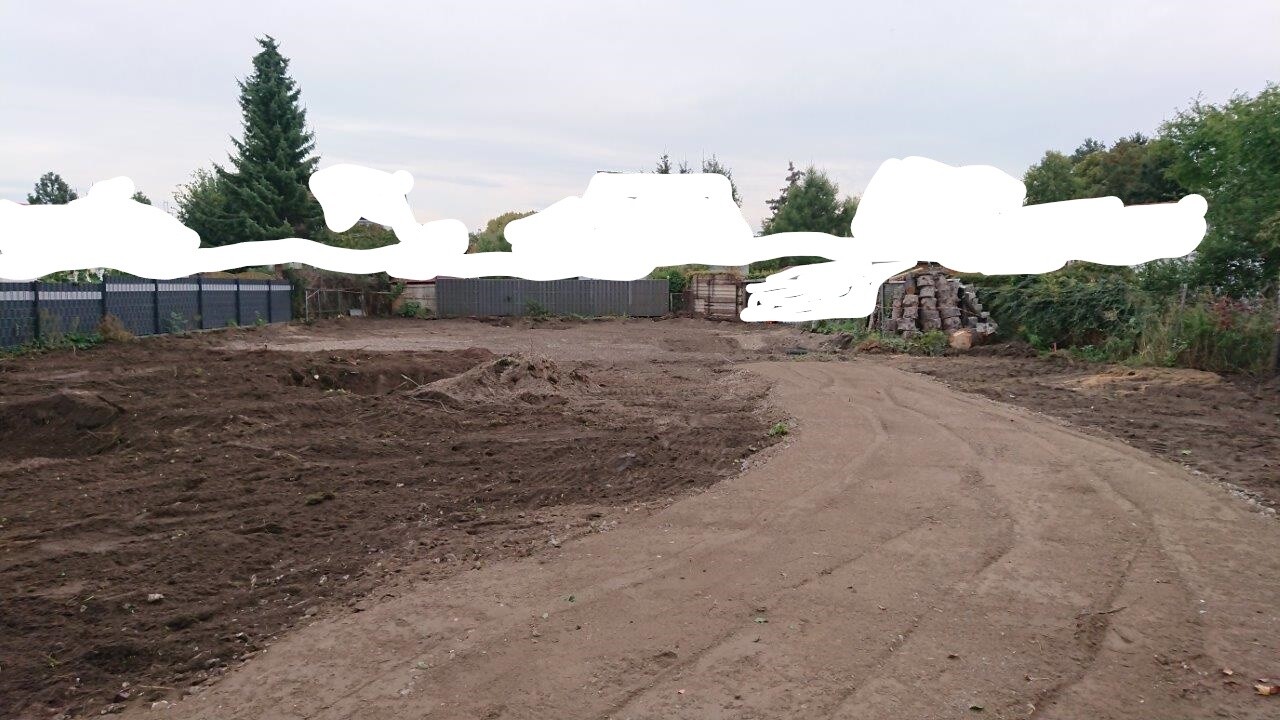 Baustelle mit unbefestigtem Weg, Erdhaufen, Holzstapel und Zaun im Hintergrund.