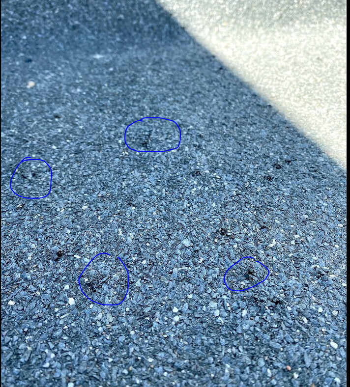 Fünf kleine dunkle Löcher in rauem Asphalt, neben einer hellen Betonkante.