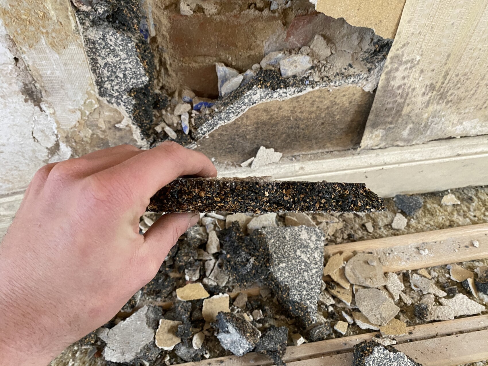 Hände halten dünnen schwarzen Asphaltstreifen vor zerbrochener Wand und Geröll.