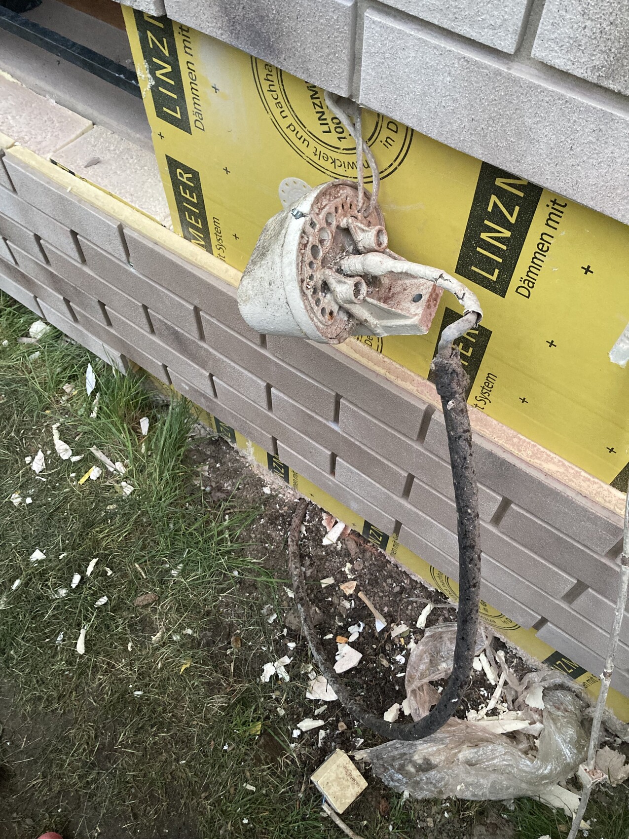 Rostiges Metallteil mit Lochscheibe hängt an Wand; Kabel und Baureste liegen daneben.