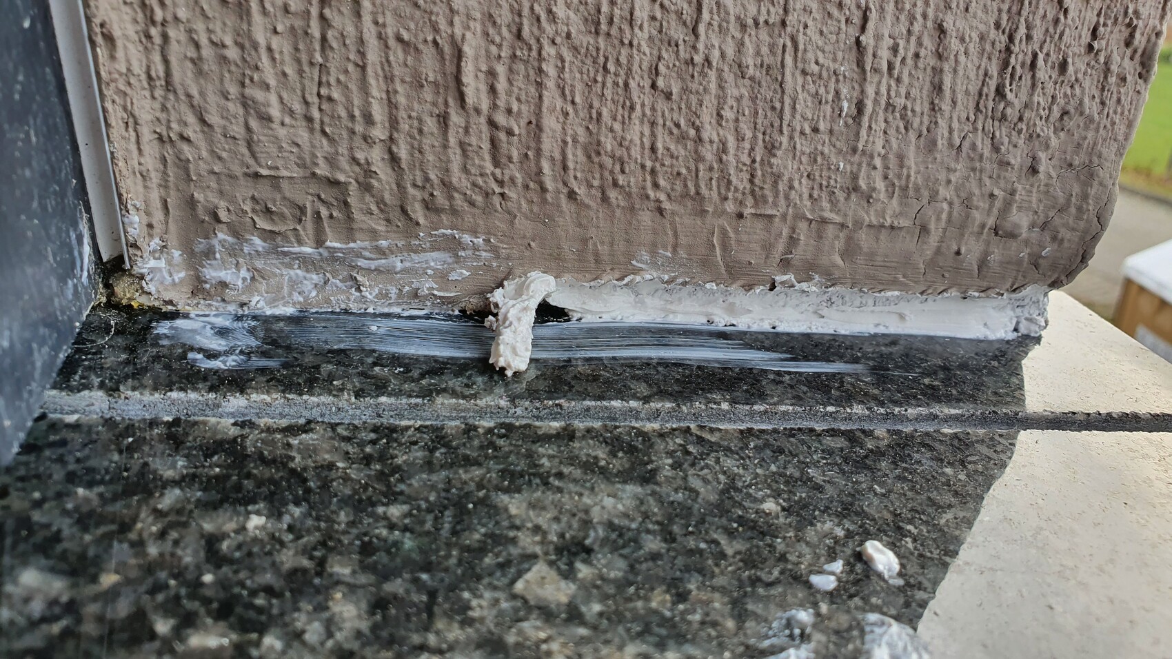 Nahaufnahme einer rauen braunen Wand mit weißer Dichtmasse an der Fuge über Steinplatte.