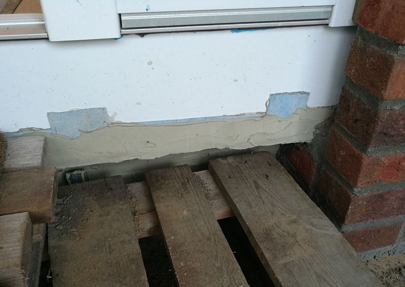 Außenwand mit abgeplatztem Putz und Holzpaletten davor, sichtbares Fundament.