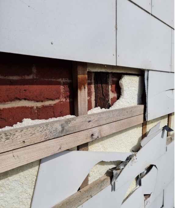 Freigelegte Backsteinwand hinter teilweise abgerissener Außenverkleidung aus Holzpanelen.