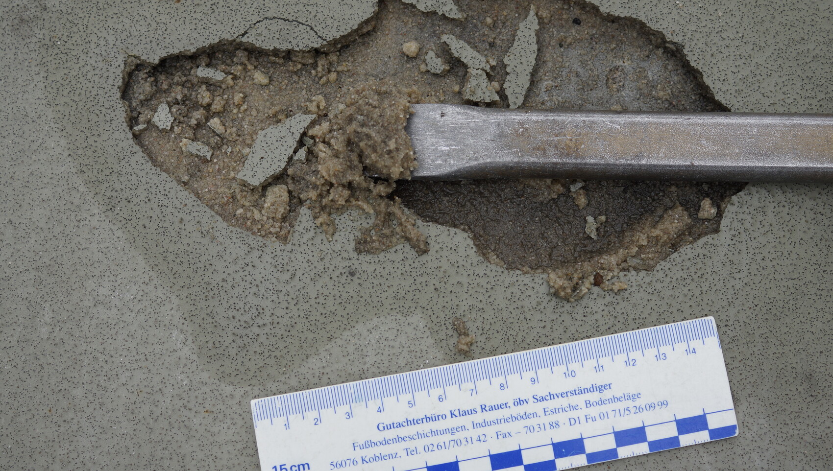 Rissiger Betonboden mit offener Bruchstelle, darüber verläuft ein Metallstab; unten Messlatte als Maßstab.