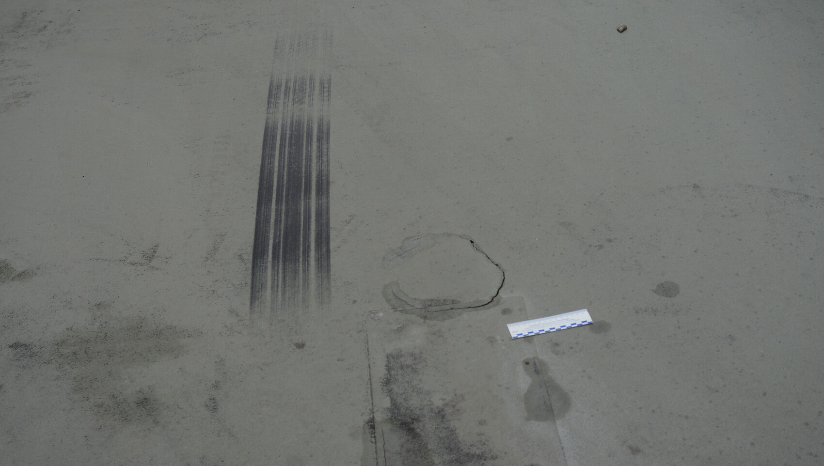 Betonboden mit langen vertikalen Reifenabdrücken, kreisrundem Riss und Messstreifen.