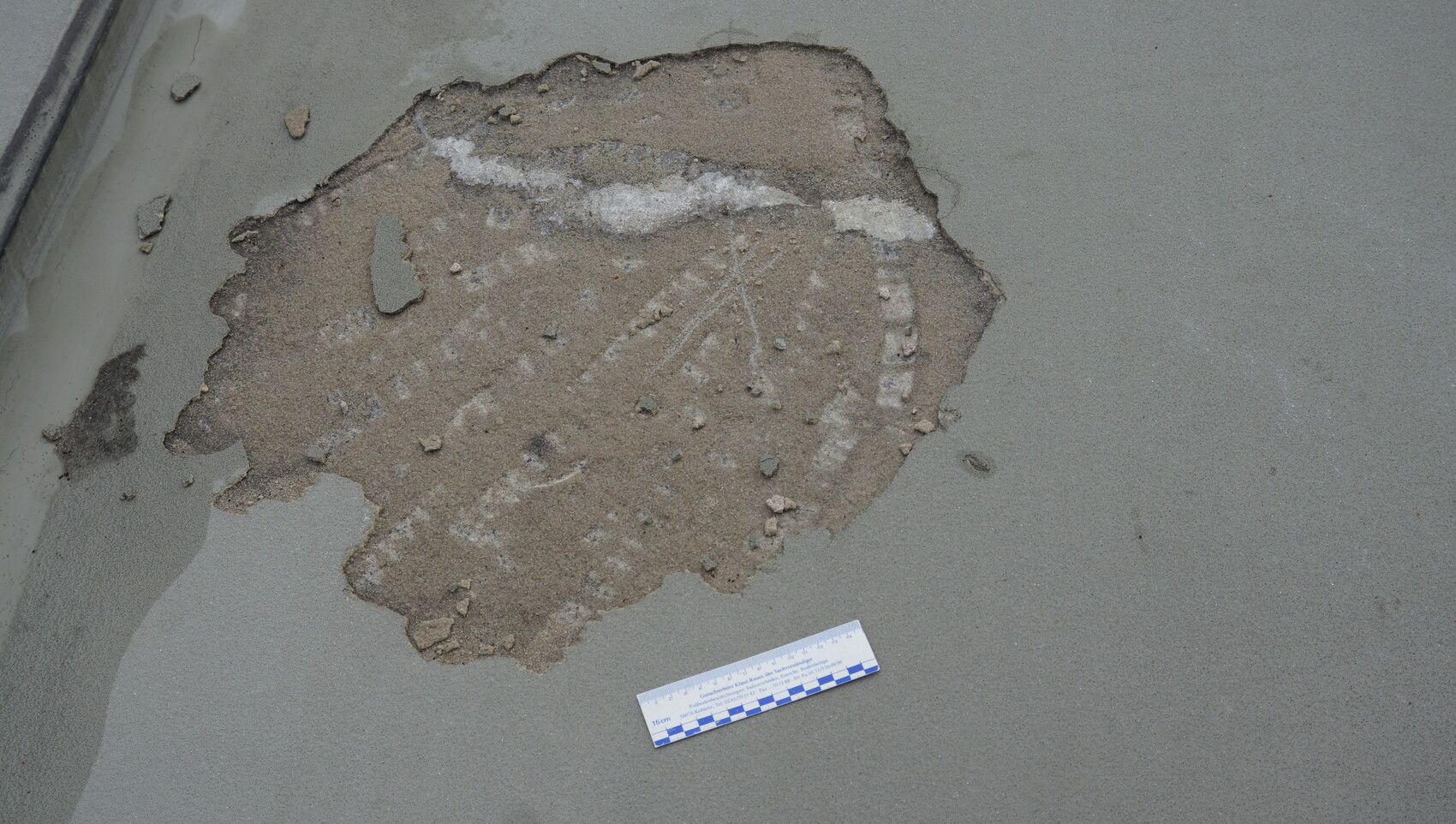 Teilweise freigelegte Erd- und Kies-Schicht in einer beschädigten Betonfläche, daneben eine Messskala.
