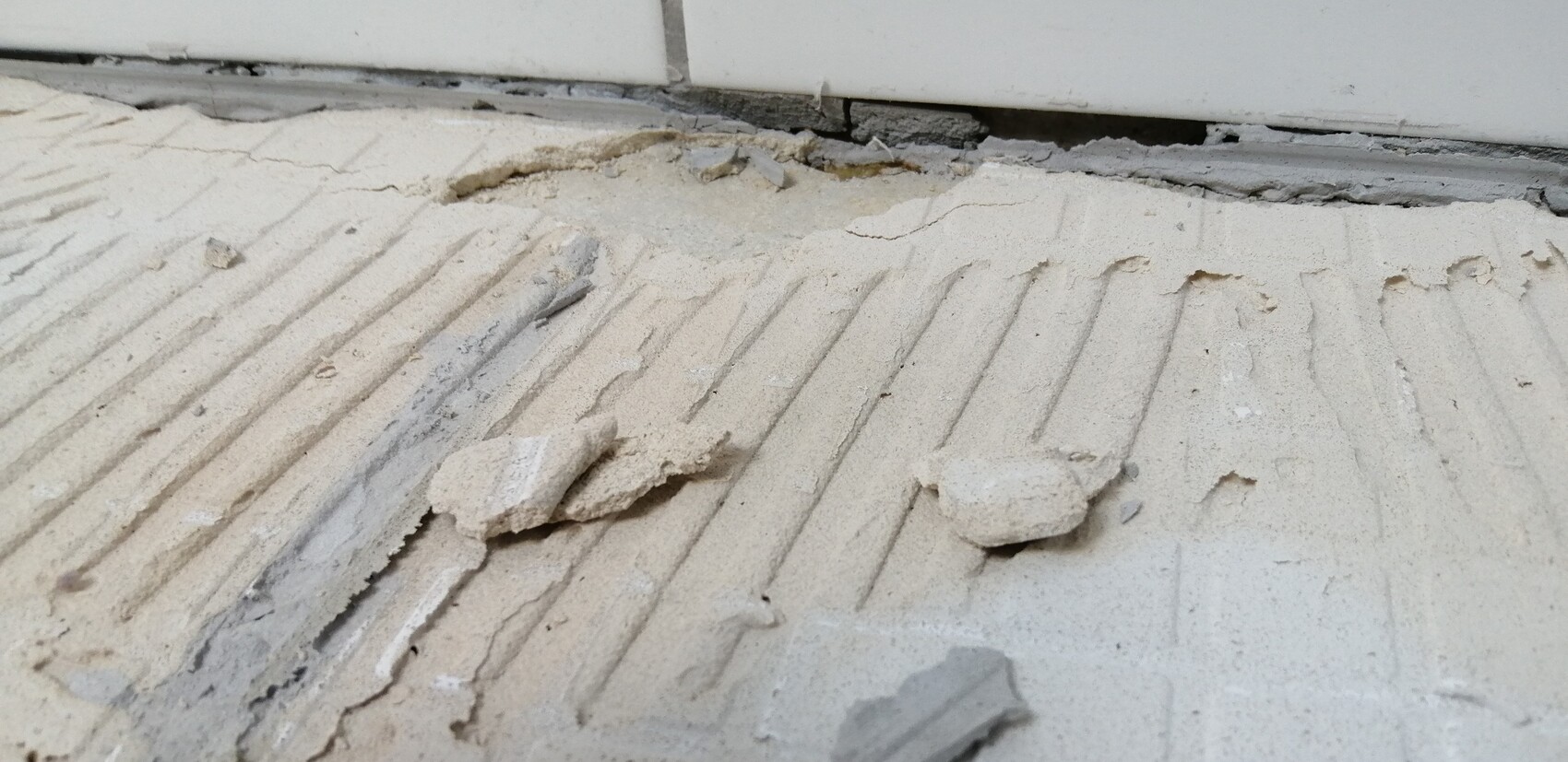 Zerbrochene Putzstücke auf einem geriffelten Estrichboden vor einer Türkante.