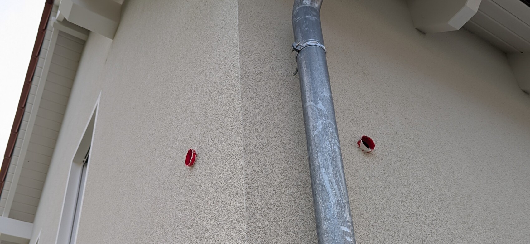 Außenwand eines Gebäudes mit Fallrohr und zwei roten Wandlöchern.