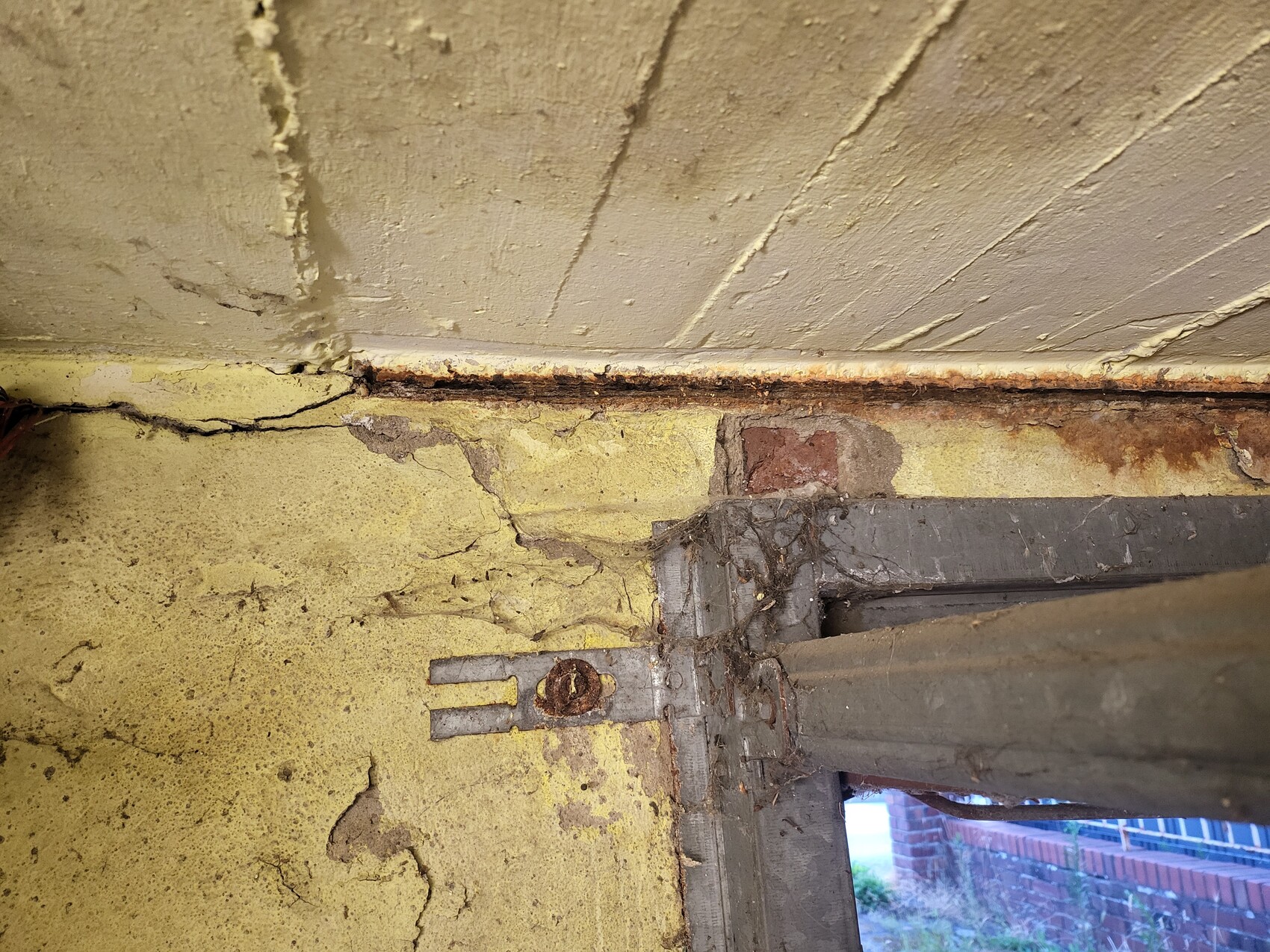 Brüchige gelbe Wand mit Rissen, Rostspuren und einer rostigen Metallverankerung am Fensterrahmen