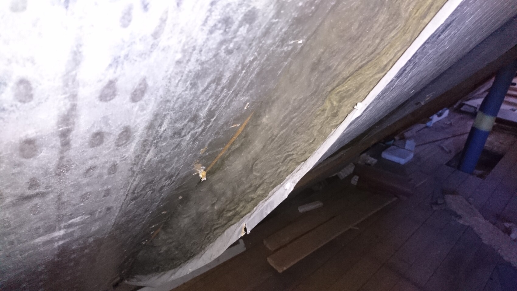 Graue Betonwand mit runden Abdruckmustern; Boden mit Holzplanken und blauer Rohrleitung.