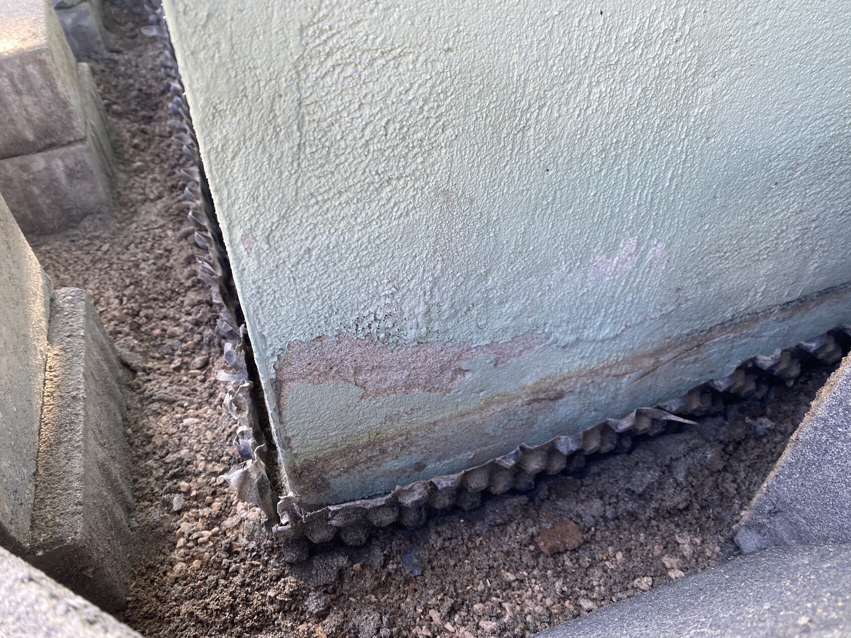 Grünlich verfärbte Betonwand mit Rostflecken am unteren Rand, Erde davor.