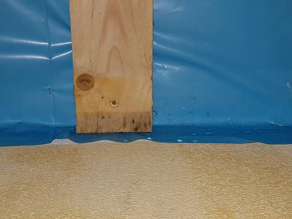 Nahaufnahme eines Holzbalkens an blauer Baufolie mit Feuchtigkeit am Boden