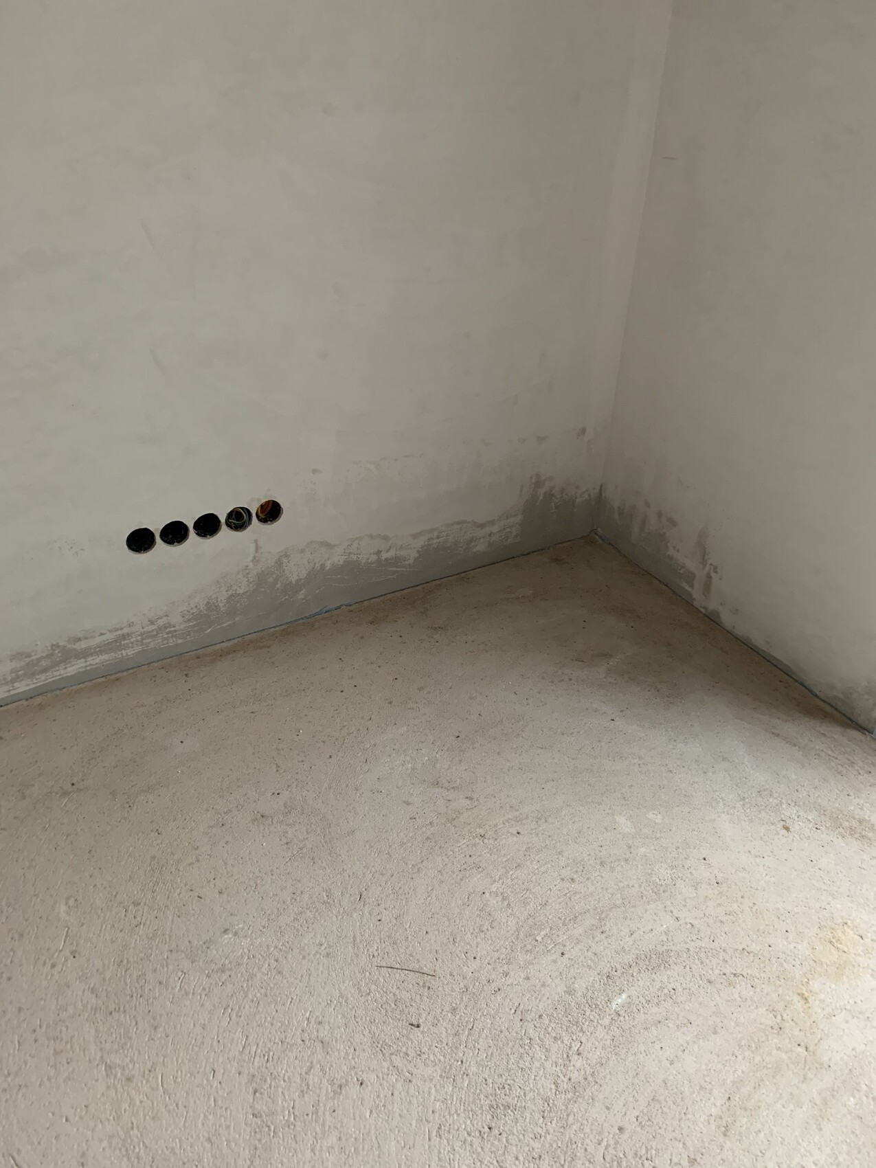 Unfertige Raumecke mit fünf runden Wandlöchern unten an der Wand; Kabel im rechten Loch sichtbar.