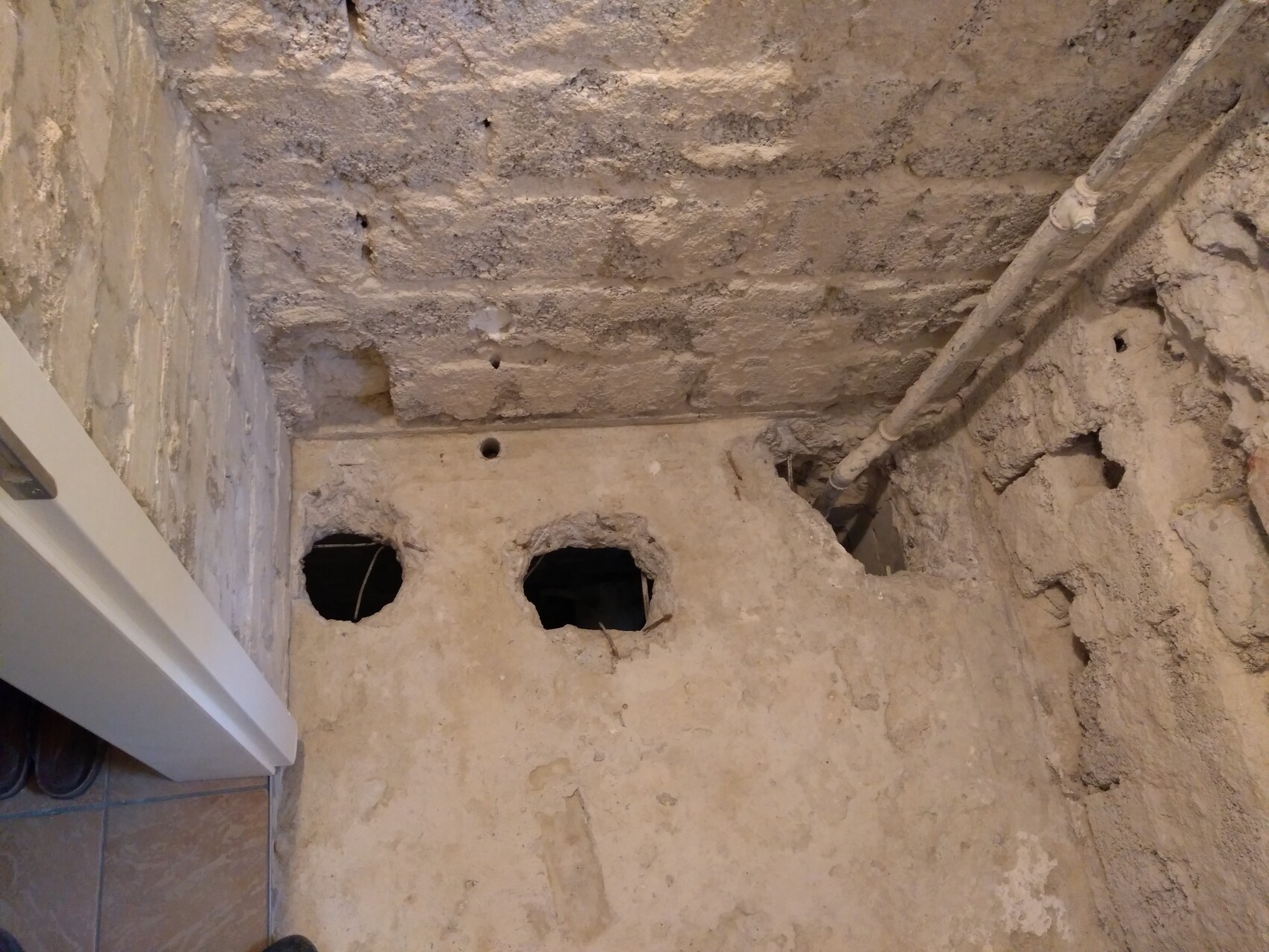Kellerboden mit zwei runden Löchern in der Betonplatte, rauer Wand und Rohren