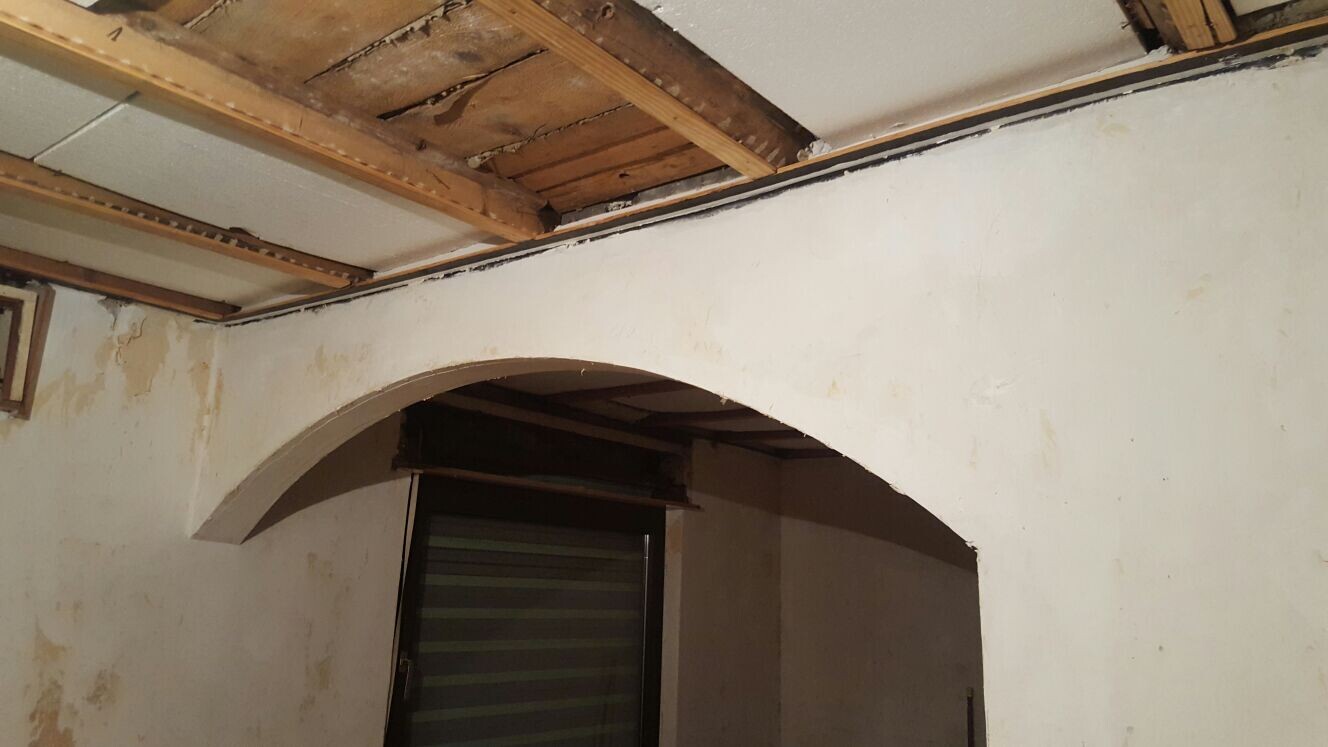 Innenraum mit freigelegten Holzbalken, Bogenöffnung und beschädigten Wänden.