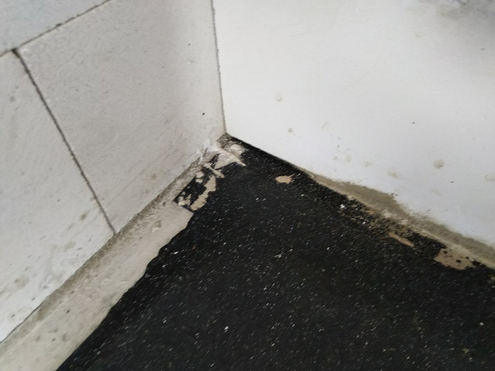 Ecke: Weiße Wand trifft graue Fliesen, dunkler Boden; Staub, Flecken und Abplatzungen am Sockel.