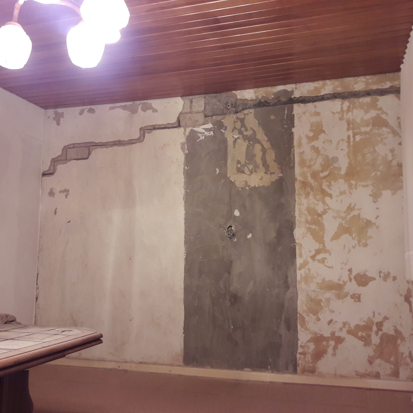 Innenraumwand mit abblätterndem Putz, dunkler Reparaturfläche und Holzdecke