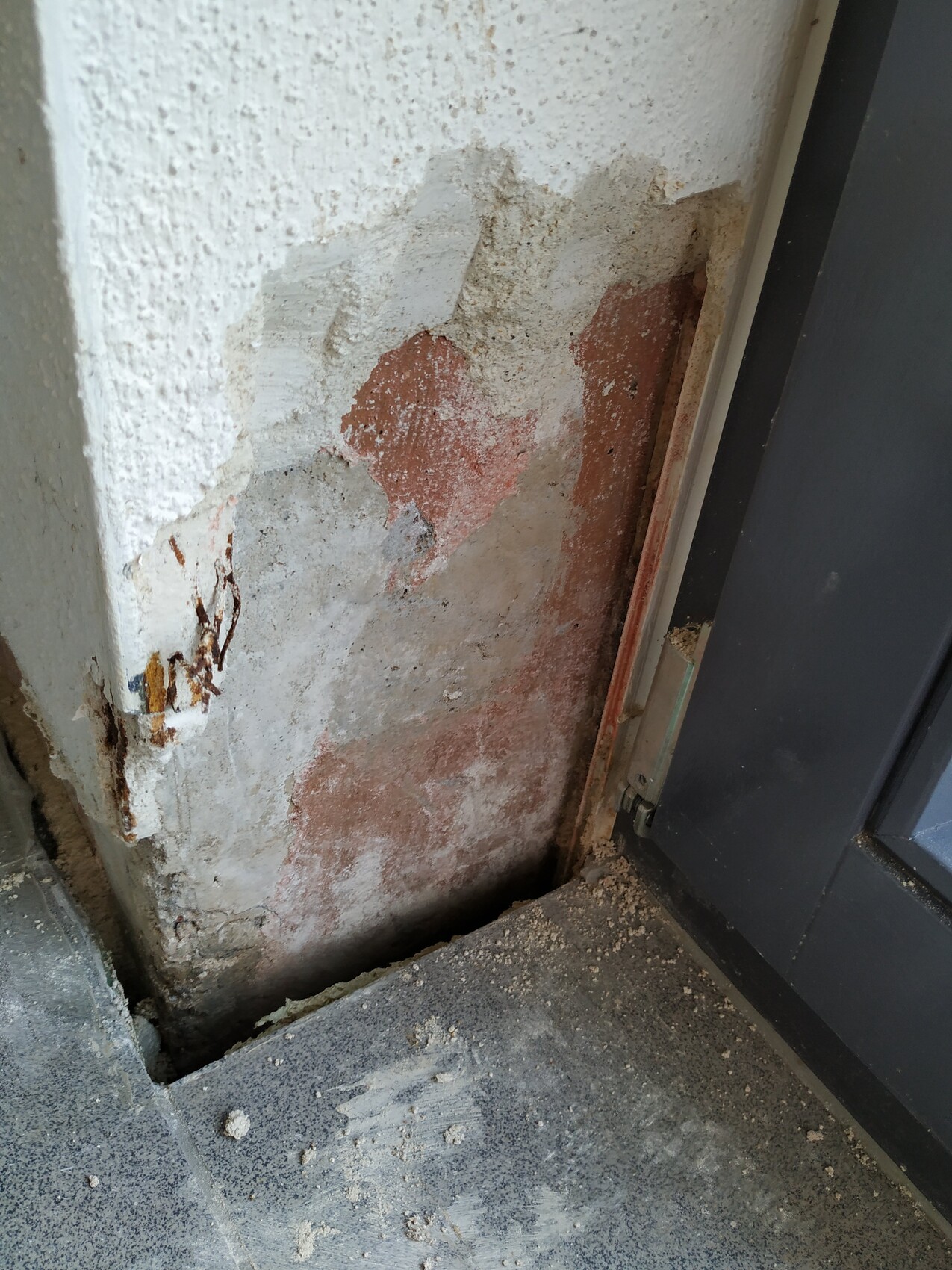 Wandbereich mit abgeplatztem Putz und rotem Ziegeluntergrund neben Eingangstür.