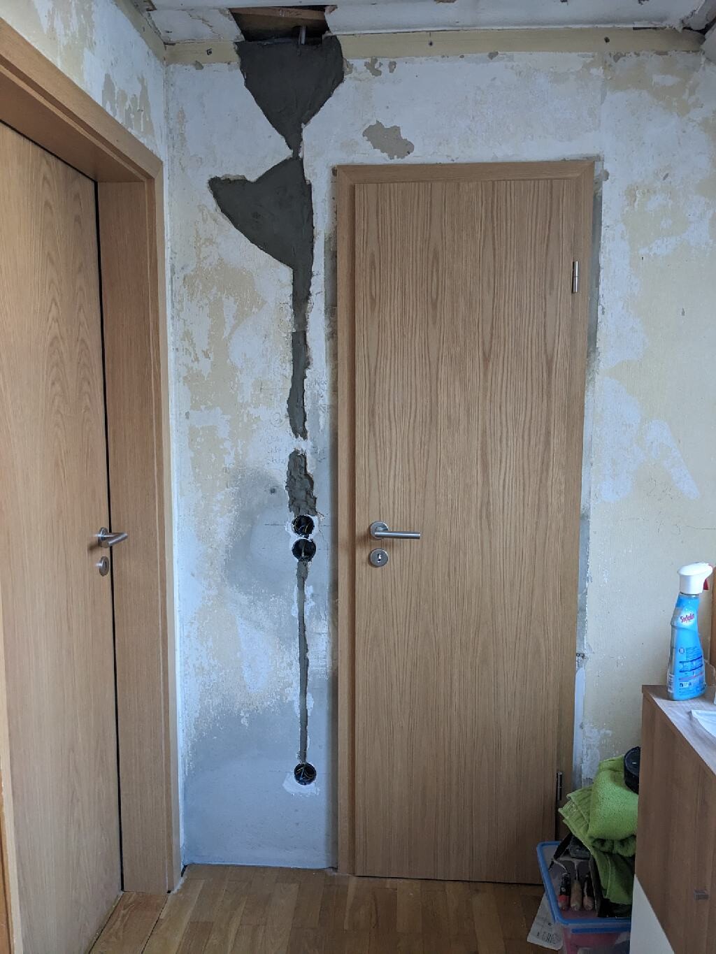 Flur mit beschädigter Wand: großer vertikaler Riss und freigelegte Öffnungen, Holztüren.