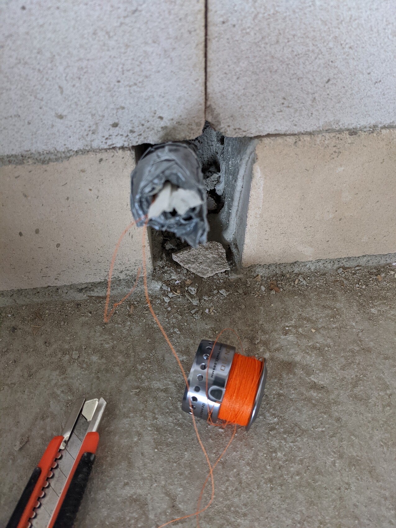Offenes Wandloch in Betonmauer, lose Leitung, orange Schnur auf Spule, Cutter daneben.